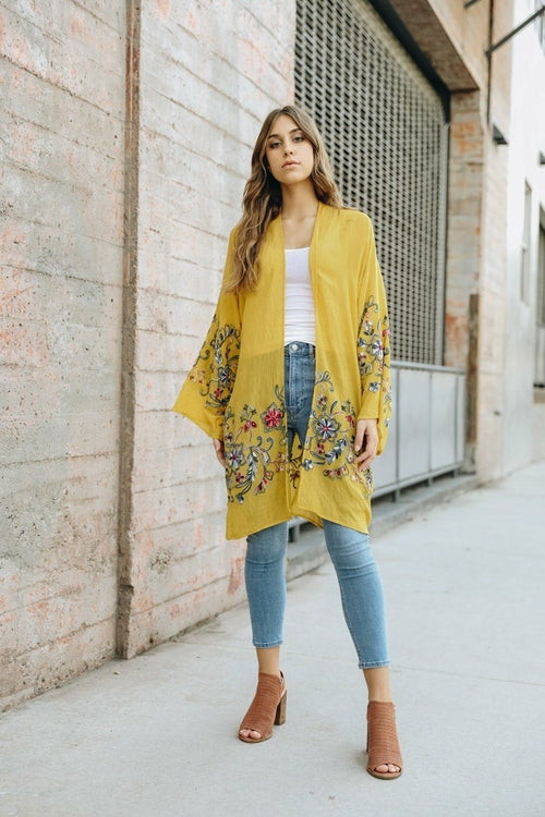 Woman wearing a yellow floral kimono over a white top and blue jeans, standing against a brick wall.