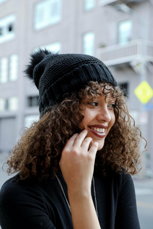 Woman wearing a black knit beanie with a pom-pom, smiling outdoors.