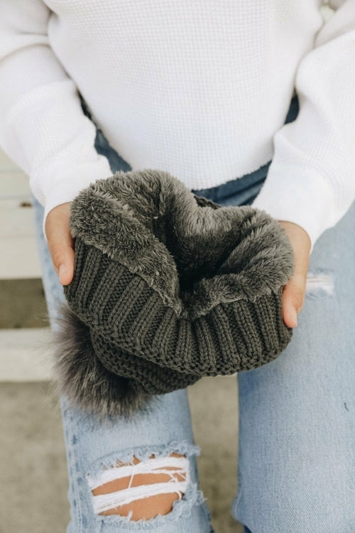 Person wearing dark gray knitted gloves with fluffy cuffs, holding them in front of a blurred background.