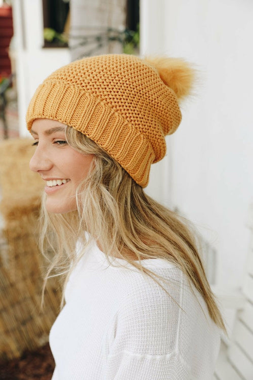 Woman wearing a mustard yellow knit beanie with a pom-pom, standing against a white wall.