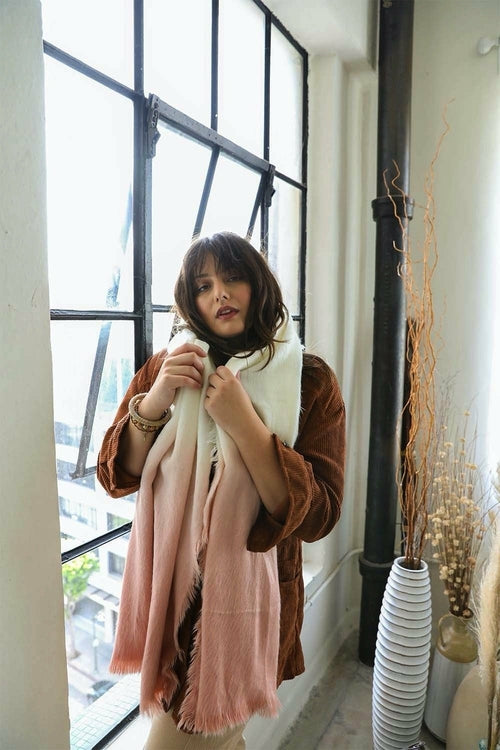 Woman holding a pink scarf in front of a large window with a vase and decorative items on a shelf.