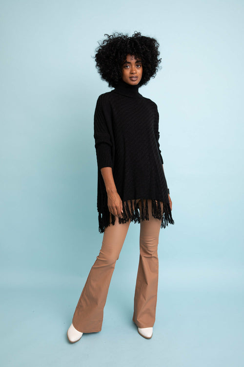Woman wearing a black turtleneck sweater with fringe and beige pants on a light blue background