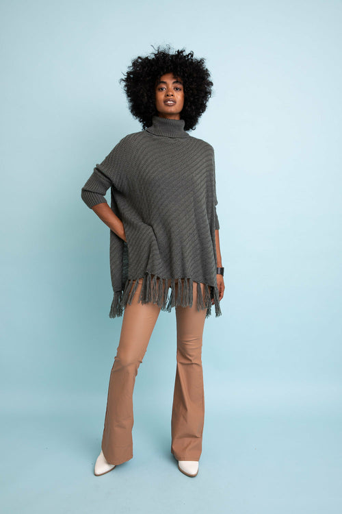 Woman wearing a gray poncho with fringe, beige pants, and white shoes on a light blue background