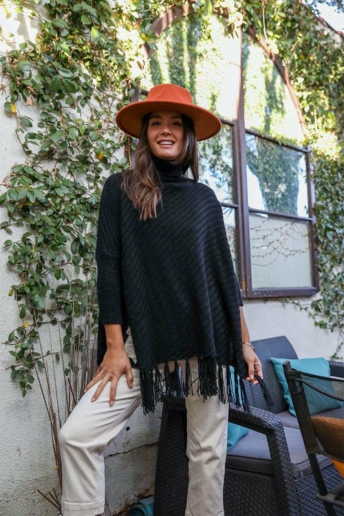 Woman wearing a black poncho with fringe, beige hat, and beige pants standing outdoors.