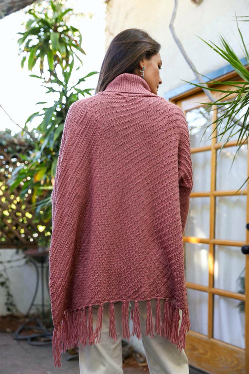 Person wearing a pink knitted poncho with fringe, standing outdoors.