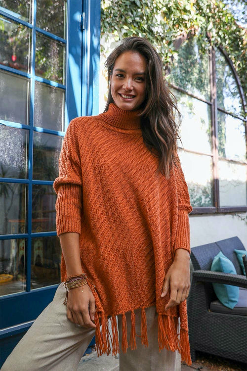Woman wearing an orange sweater with fringe standing outdoors.