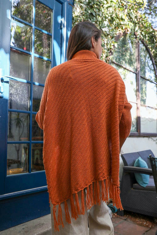 Person wearing an orange knitted cardigan with fringe, standing in front of a blue door.