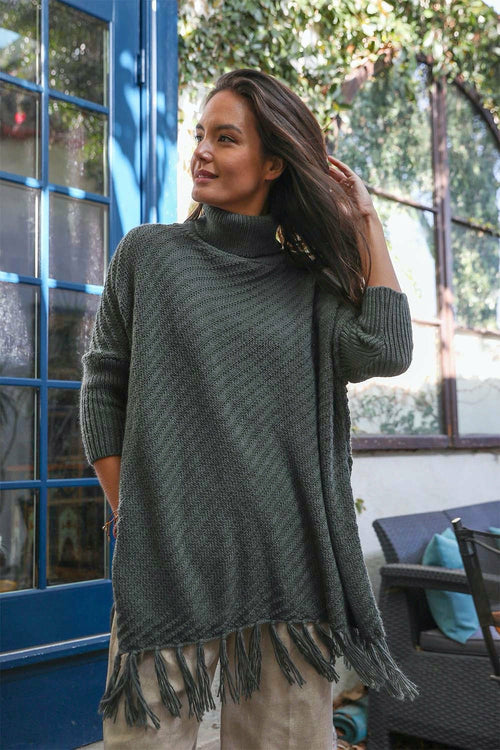 Woman wearing a green poncho with fringe standing outdoors near a building.