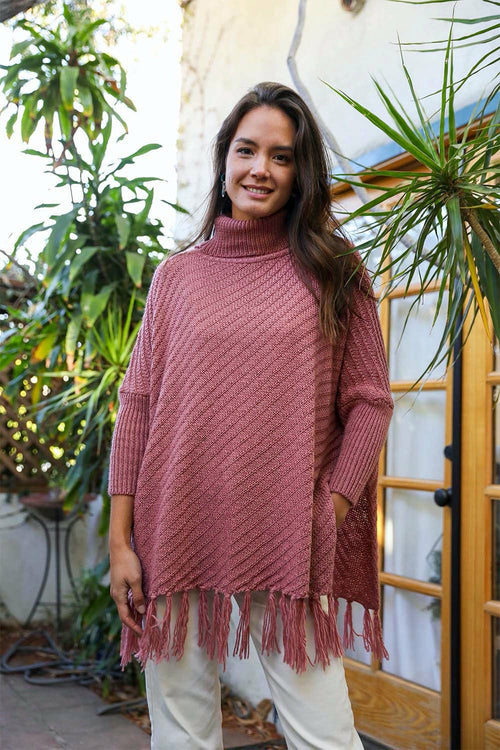 Woman wearing a pink knitted poncho with fringe standing outdoors near plants.