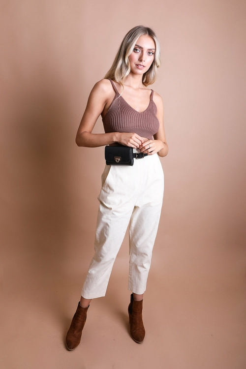 Woman wearing a brown tank top, white pants, and brown boots against a beige background