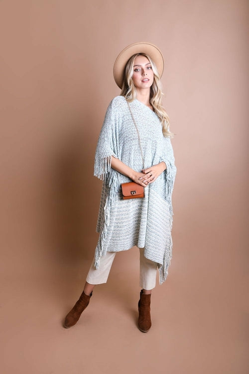 Woman wearing a light blue poncho with fringe, beige hat, white pants, and brown boots against a beige background.
