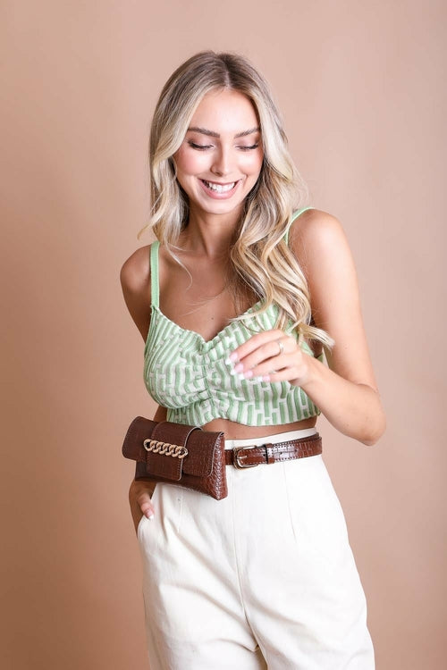 Woman wearing a green top and white pants with a brown belt against a beige background