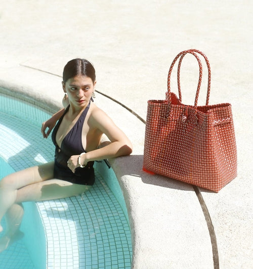Woman in a swimsuit sitting by a pool with a woven bag next to her