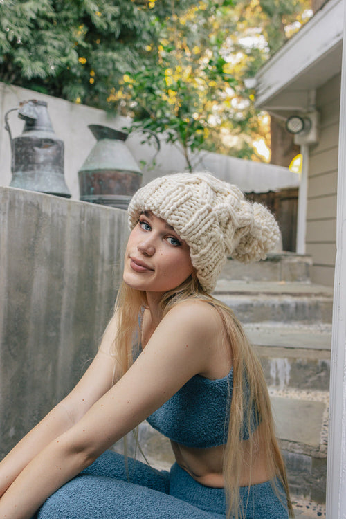Woman wearing a beige knit hat sitting outdoors with a house and trees in the background