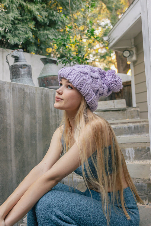 Woman wearing a purple knitted beanie sitting outdoors with trees and a house in the background.