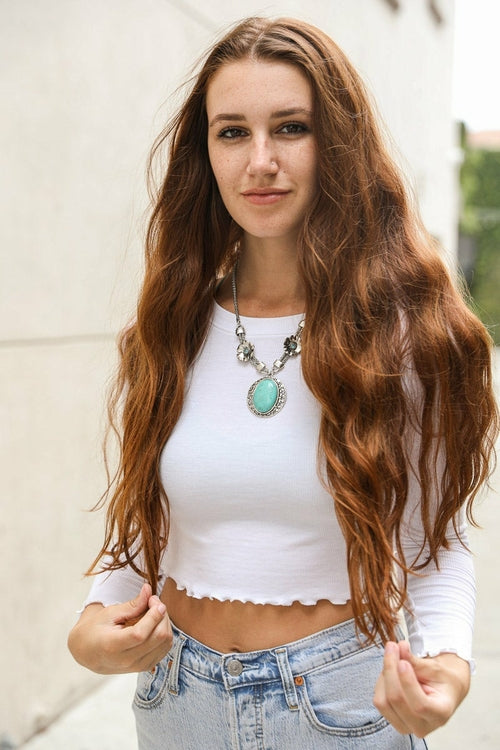 Woman with long brown hair wearing a white top and blue jeans, standing against a light-colored wall.