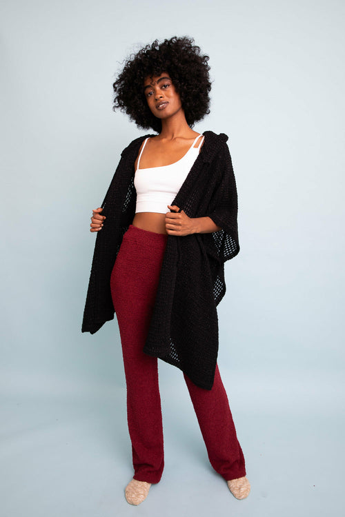 Woman wearing a black cardigan, white top, and red pants on a light gray background