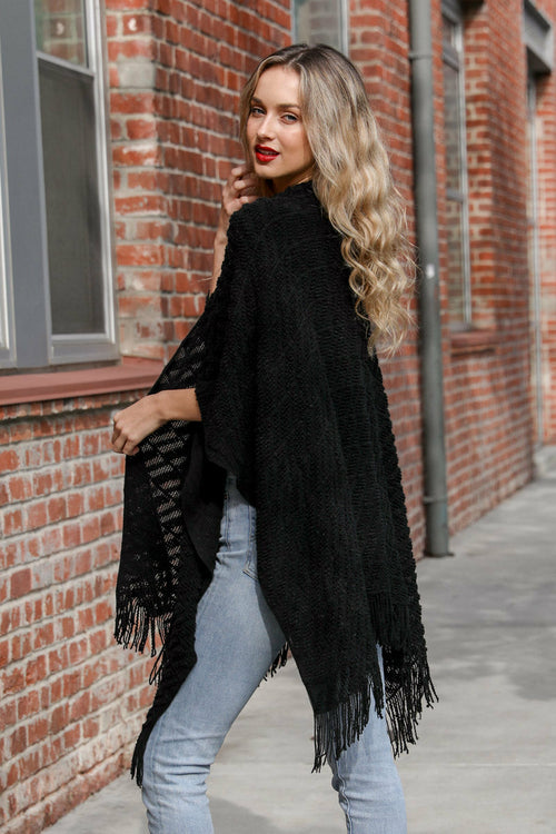Woman wearing a black fringed poncho against a brick wall.
