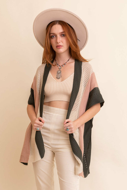 Woman wearing a beige hat, beige top, and pants with a pink and black cardigan against a beige background