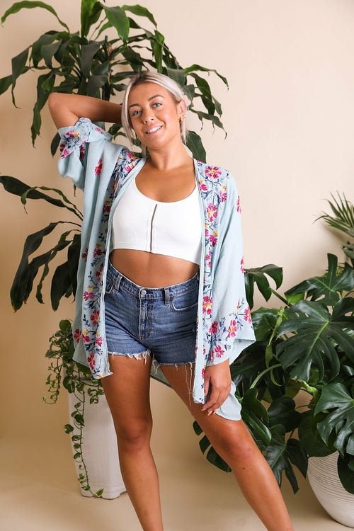 Woman wearing a floral kimono over a white crop top and denim shorts, standing in front of green plants.
