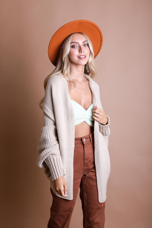 Woman wearing a beige cardigan, white top, brown pants, and orange hat against a beige background