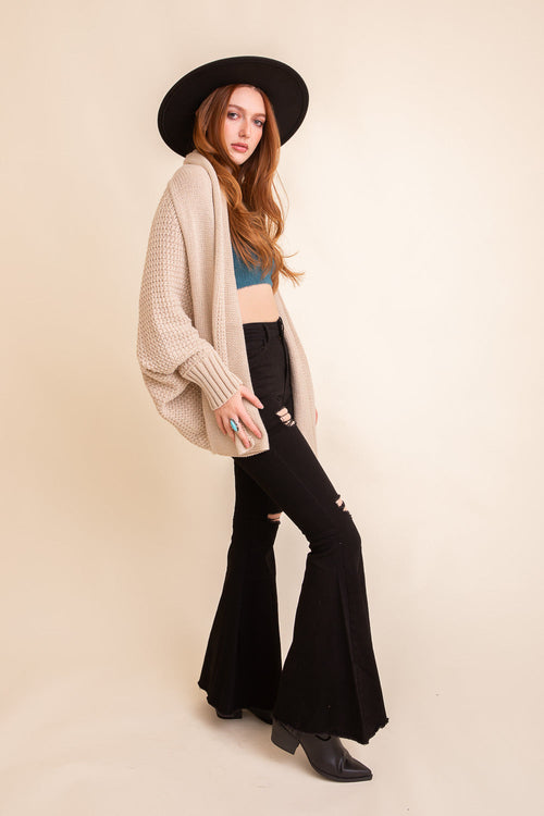 Woman wearing a beige cardigan, black top, and black flared pants with a black hat on a beige background