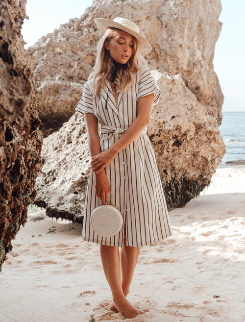 Woman in a striped dress and hat standing on a beach with large rocks and ocean in the background