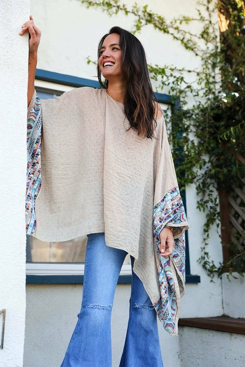 Woman wearing a beige poncho with floral sleeves in front of a white wall with greenery.
